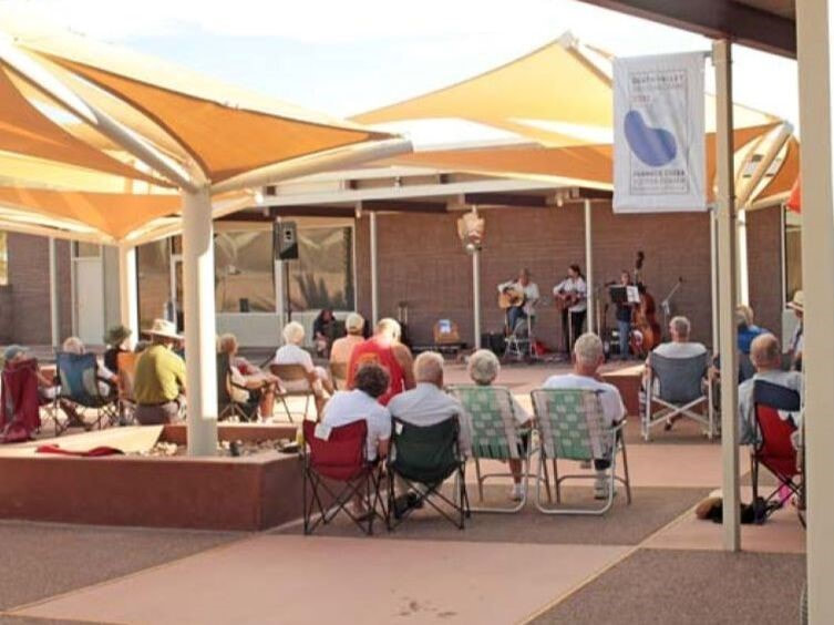 A group of people sit in folding chairs underneath triangular shade canopies watching a trio of musicians