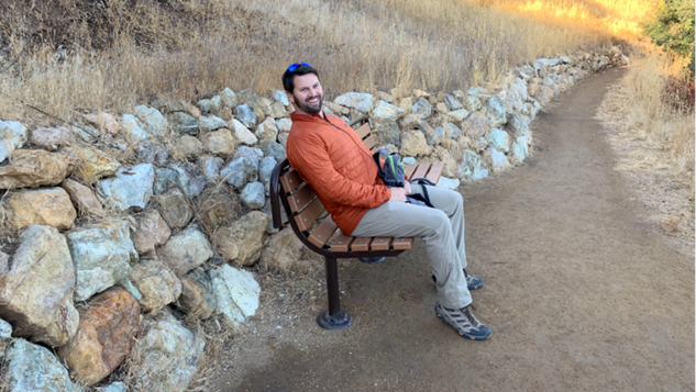 A person sits on a bench in front of a stone wall along a dirt trail