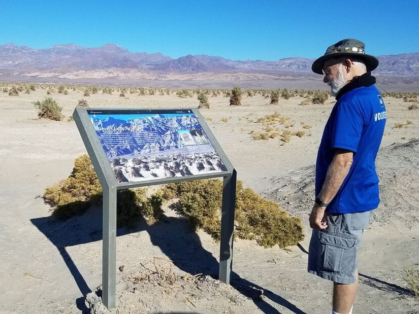 A visitor looks at an educational sign with sand dunes and mountains in the background