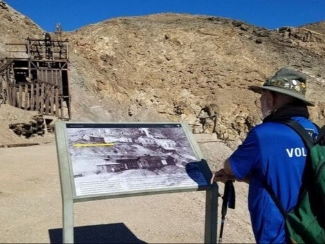 A person stands reading an educational sign in front of a large wooden mining structure and a rocky mountainside