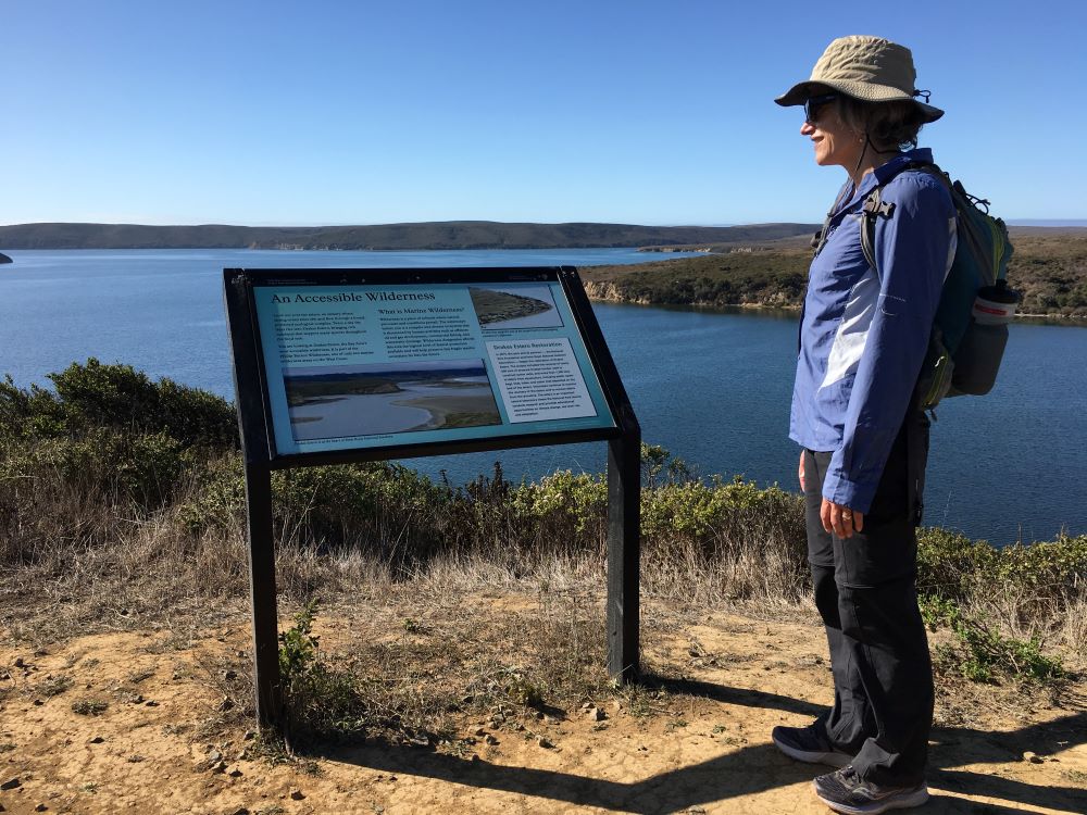 A person stands looking at an educational sign while standing on a bluff overlooking the ocean