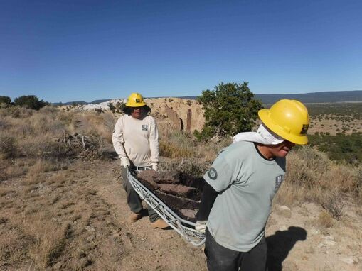 2 Ancestral Lands Conservation Corps members haul asphalt on a metal litter on a trail along the edge of a bluff overlooking desert plains.
