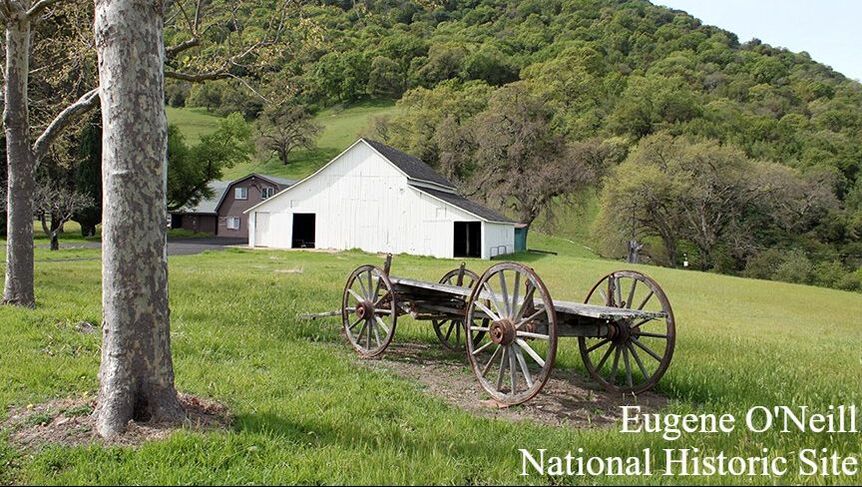 The base and wheels of a historic wagon sits in a grassy area in front of a large white barn with oak trees on a hillside behind