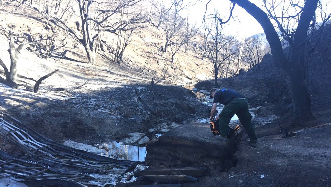 An NPS ranger leans over a backpack on a bridge abutment next to a charred landscape. Pieces of an old bridge lay flat across a small creek.
