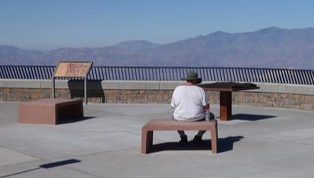 A person sits at a scenic viewpoint on a red cement bench facing away from the camera; an educational sign, bronze map, and mountain range are in the background