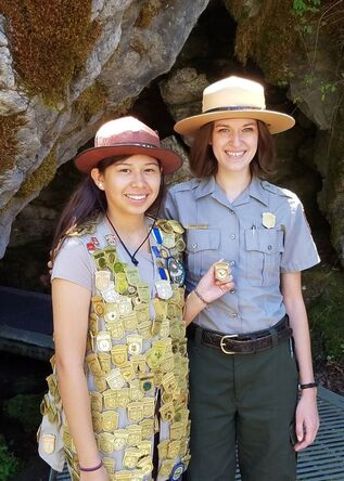 A junior ranger wearing a vest full of junior ranger badges and holding an Oregon Caves badge, stands with a National Park ranger in front of a cave opening