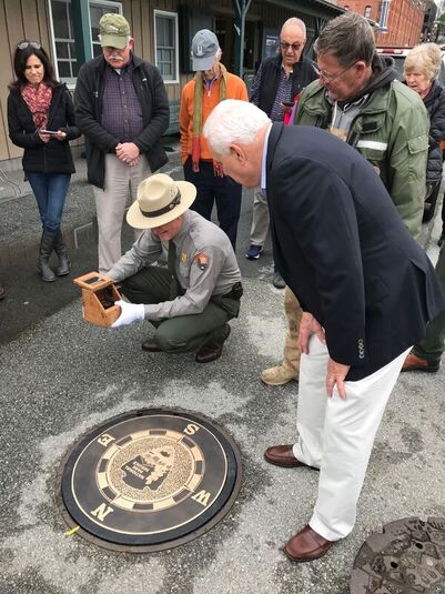 A National Park Service ranger holds a compass in a small wooden box and peers at the compass rose inlay to orient it while a group of people looks on 