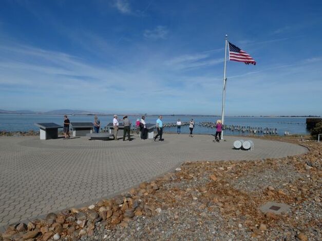 People walk about at Port Chicago, a paved open area adjacent to the ocean with wayside signs and an American flag