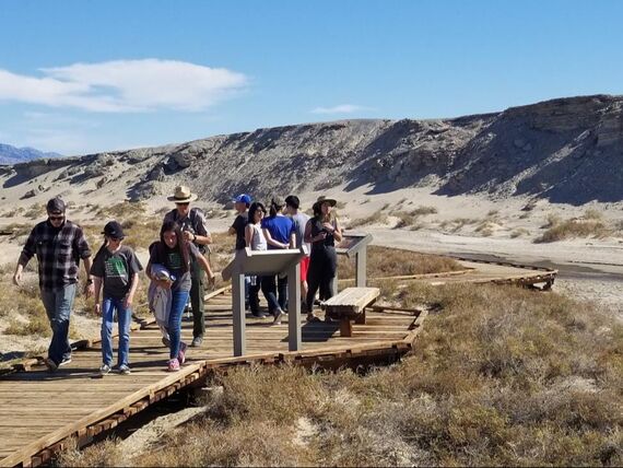 A group of students with National Park Service rangers stands and walks on a boardwalk in front of two educational signs