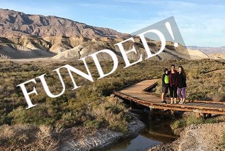 A group of three people stand on a wooden boardwalk above a shallow creek in the desert