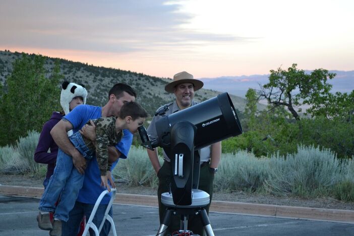 A visitor holds up a young child to look through a large telescope while a ranger looks on