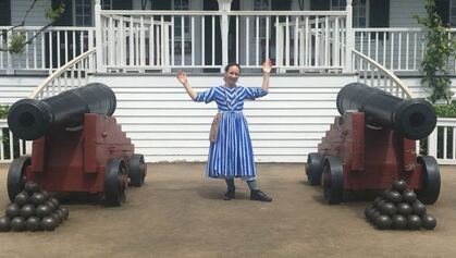 A person in a blue and white dress stands in front of the steps of a large house and between two large cannons, each with a pyramid stack of cannonballs in front of them