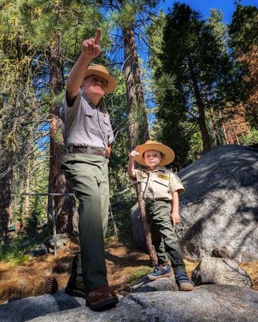 A National Park Service ranger in uniform stands pointing in the distance next to a small Junior Ranger, also in uniform and pointing in the same direction