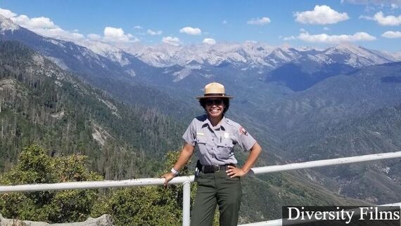 A ranger in uniform poses in front of a railing with a large mountain landscape in the background