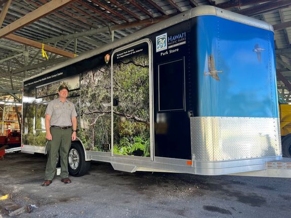 A National Park Service ranger in uniform stands next to a large trailer that has tropical vegetation imprinted on the outside and the words 
