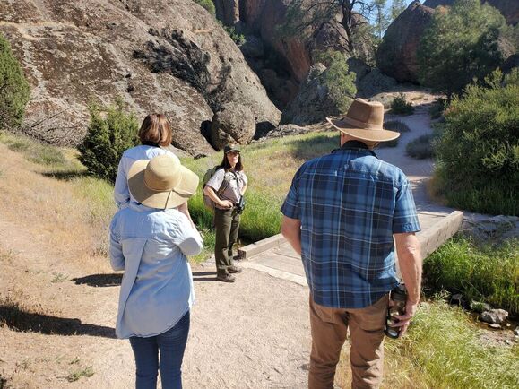 A National Park Service ranger stands talking with a group of 3 people near a bridge set among granite rocks and green vegetation