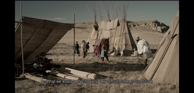 A screenshot from the park orientation film, showing people milling about traditional tule reed structures