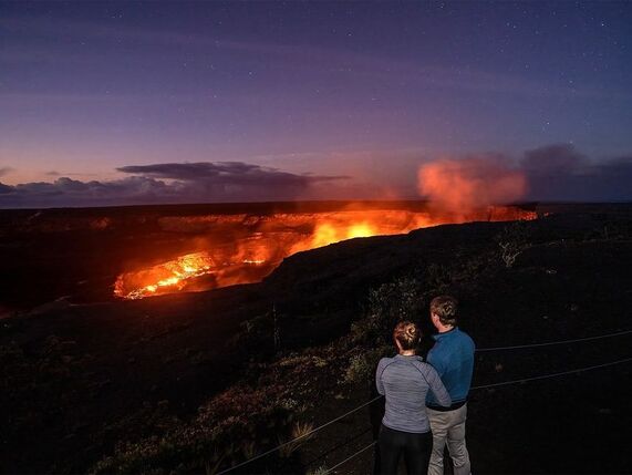 Two people stand arm in arm watching red glowing lava and steam coming off a dark volcanic landscape