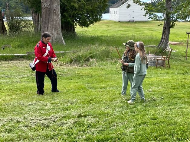 A person in a replica historic British military uniform holds a wooden hoop and stick facing two children, who also have a wooden hoop and stick, as if being taught a game