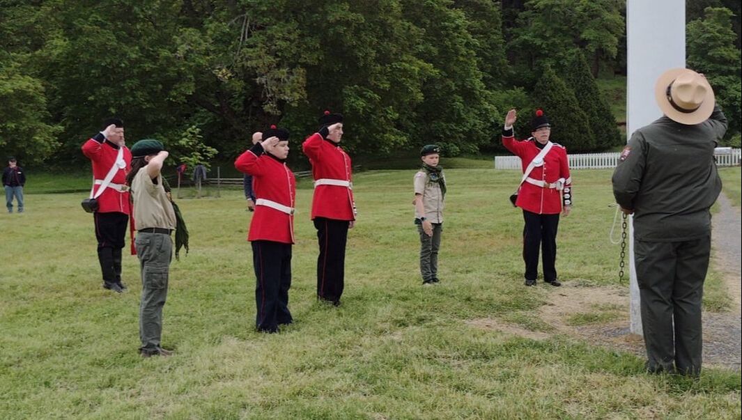 A group of individuals in historic military uniforms stands facing a flagpole and saluting. An NPS ranger in uniform is facing the group with their back to the camera