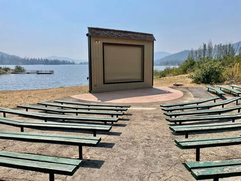 A small amphitheater made up of benches with chipped green paint and a small audiovisual kiosk and pavilion with a large blue lake and mountains in the background