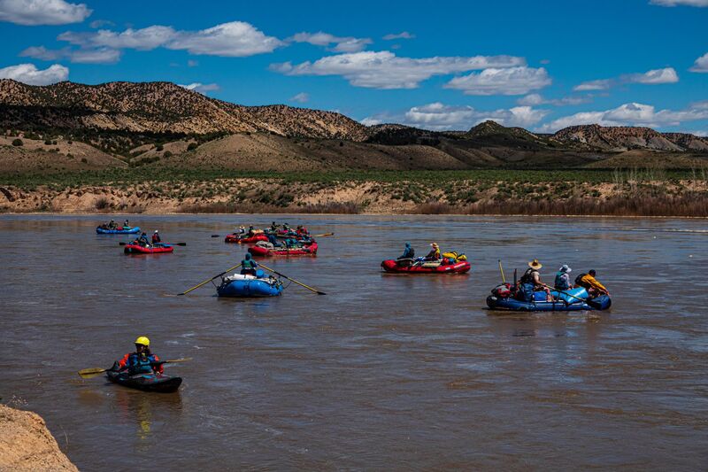 Several rafts and a kayak float down a large river along rolling brown and green hills