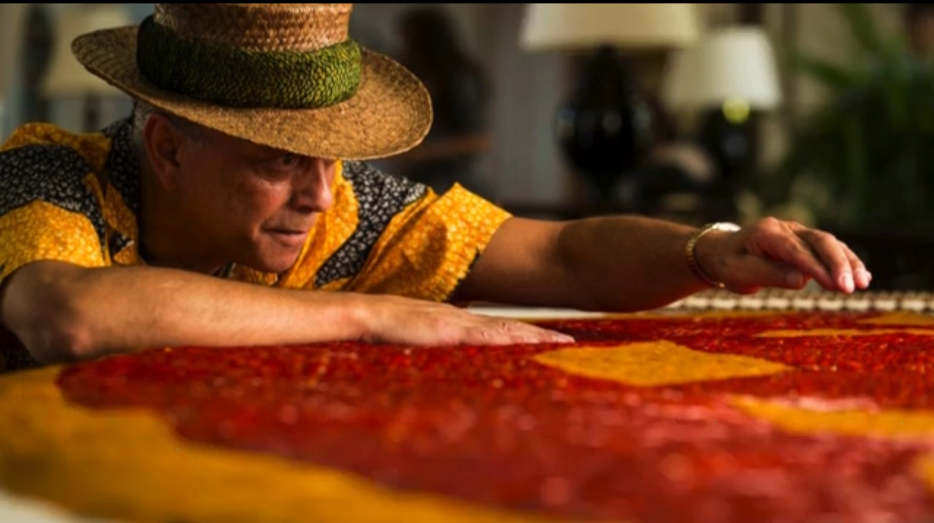 An individual wearing a banded hat leans over a red and yellow textile made from featheres