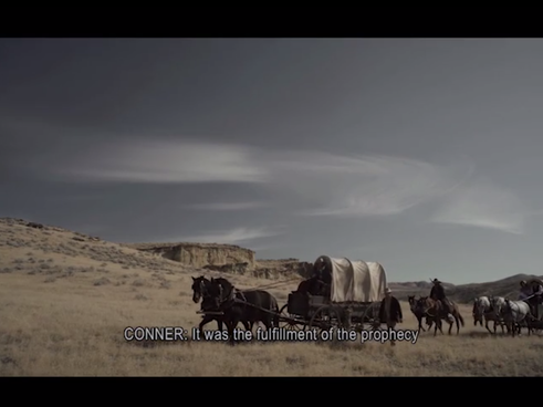 A horse-drawn covered wagon train travels across an ope plain with cliffs in the background; there is a black bar across the top indicating the borders of a film screen