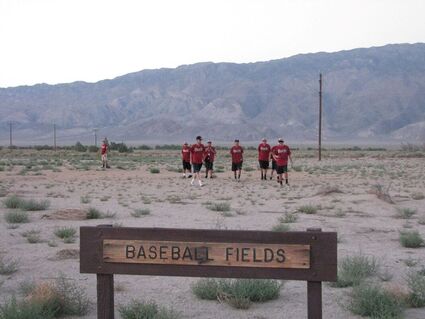 A group of people in matching red and black sports uniforms walk across a field with patchy grass behind a sign that says "Baseball Fields"