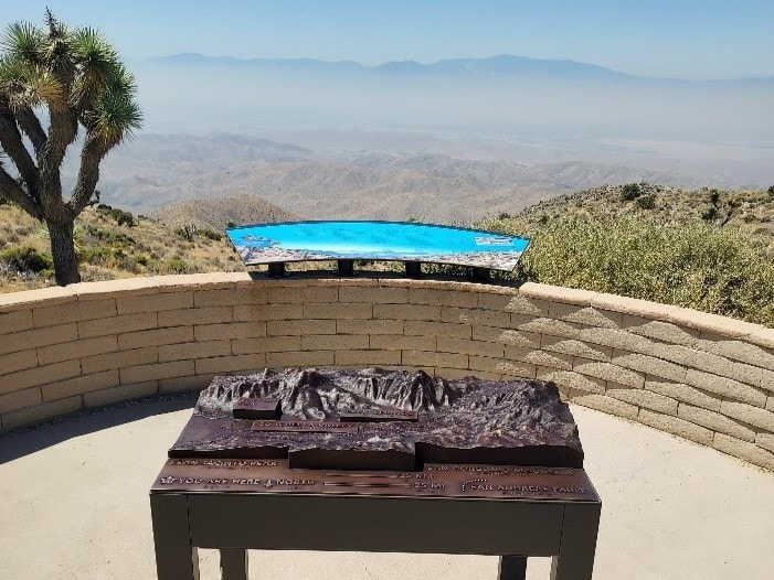 A tactile relief map sits in front of a round retaining wall with a long educational sign that shows the valley and mountains in the background