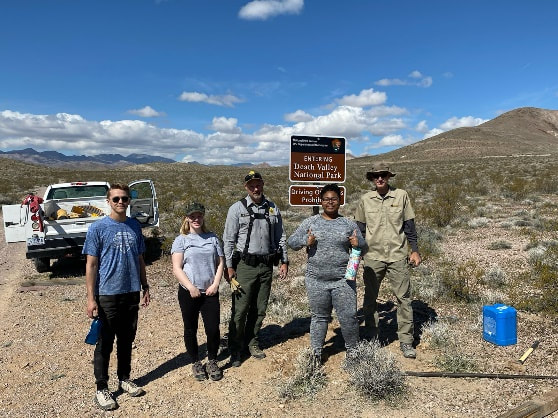 3 college student volunteers stand next to two National Park rangers and newly-installed entrance point signs with rocky mountains in the background