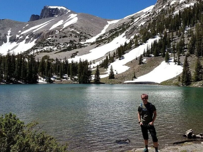 A person stands in front of an aquamarine lake against snow-dappled mountains.