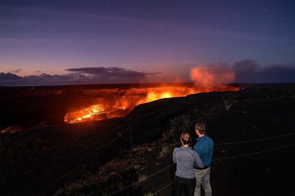 Two people stand arm-in-arm watching distant volcanic activity at night