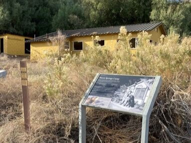 An educational sign with the title "Warm Springs Camp" sits in front of a yellow building and some vegetation